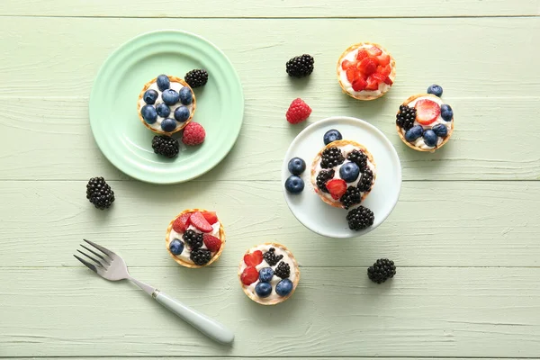 Plates of tasty tartlets with whipped cream and berries on green wooden background