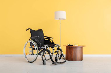 Boş tekerlekli sandalye, lamba ve sarı duvarın yanındaki masa.