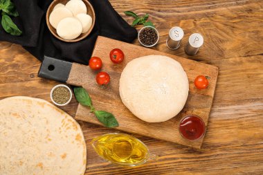 Board with raw dough and ingredients for preparing pizza on wooden background