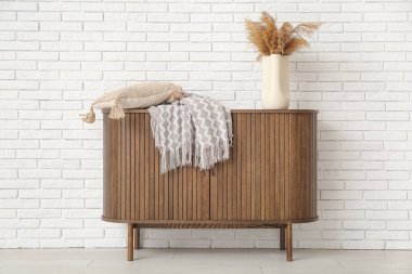 Wooden cabinet with blanket and cushion near white brick wall