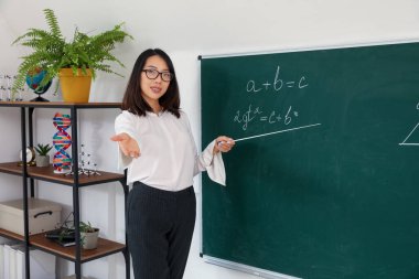 Sınıfta yazı tahtasının yanında matematik dersi veren Asyalı kadın öğretmen.