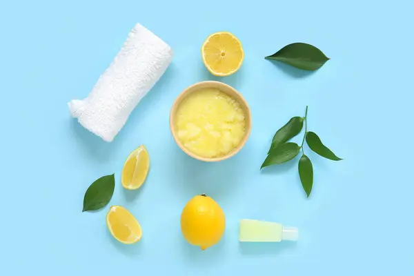 Bowl of lemon body scrub with cosmetic bottle and towel on blue background