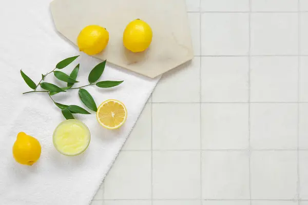 Bowl of lemon body scrub with board and towel on white tile background