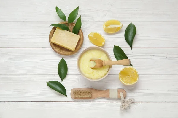 Bowl of lemon body scrub with massage brush and soap on white wooden background