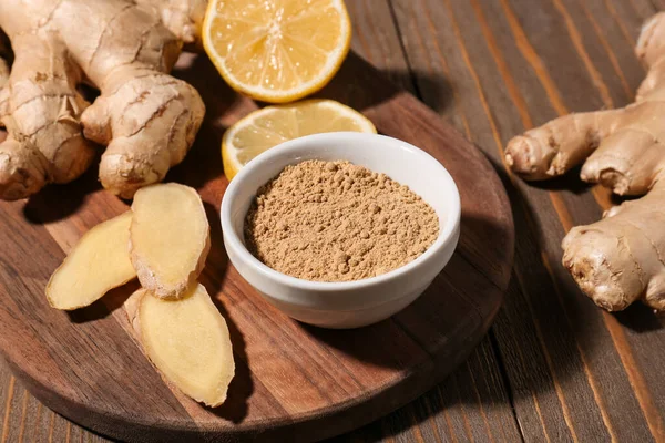 stock image Board with fresh ginger roots, lemon and bowl of dried powder on wooden background