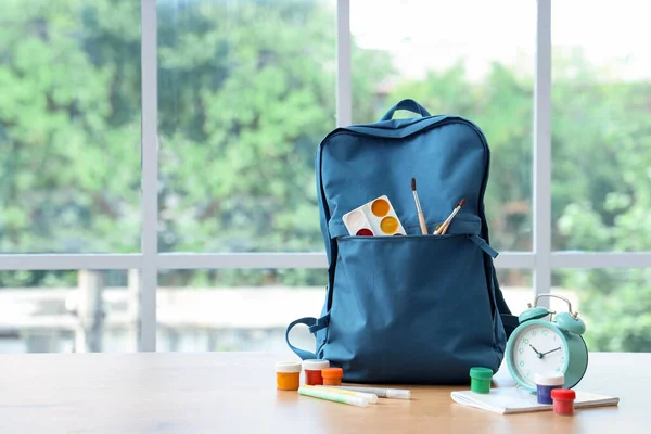 教室窗户附近的木制桌子上 蓝色校服背包 装有文具和闹钟 — 图库照片