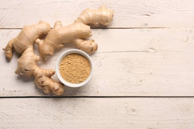 Taze zencefil kökü ve beyaz ahşap arka planda kurutulmuş toz dolu kase.
