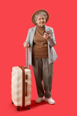 Kırmızı arka planda cep telefonu ve bavulu olan yaşlı bir kadın.
