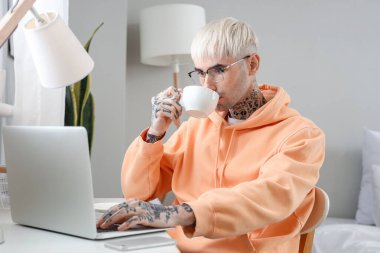 Gözlüklü, dizüstü bilgisayarlı, kahve içen genç bir adam.