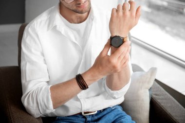 Şık bir genç adam koltukta oturuyor ve odadaki kol saatine bakıyor.