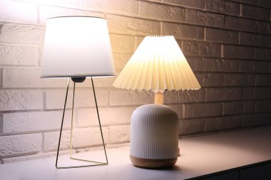 Karanlık odada beyaz tuğla duvarın yanındaki masada farklı lambalar var.