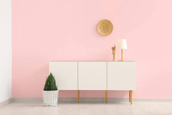 Modern chest of drawers with houseplant and plate hanging on pink wall in room