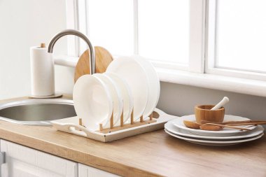 Mutfak tezgahında lavabo, tabak rafı ve pencerenin yanında temiz tabaklar var.