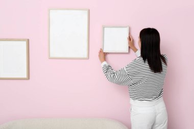 Genç bir kadın oturma odasındaki pembe duvara boş bir çerçeve asıyor.