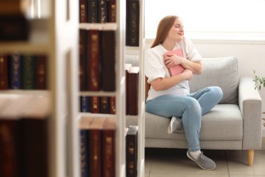 Kitaplı genç bir kadın kütüphanede koltukta oturuyor.