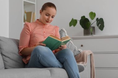 Evdeki kanepede kitap okuyan genç bir kadın.