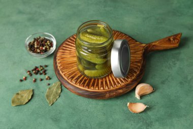 Lezzetli mayalanmış salatalıklı kavanoz, ahşap tahta ve yeşil arka planda malzemeler.