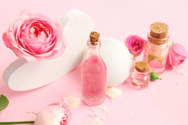 Gül özlü kozmetik yağı şişeleri ve pembe arka planda çiçekler.