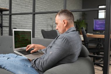 Modern dizüstü bilgisayarı olan olgun bir erkek programcı koltukta oturuyor.