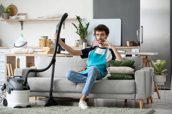 Young man with vacuum cleaner sitting on sofa at home