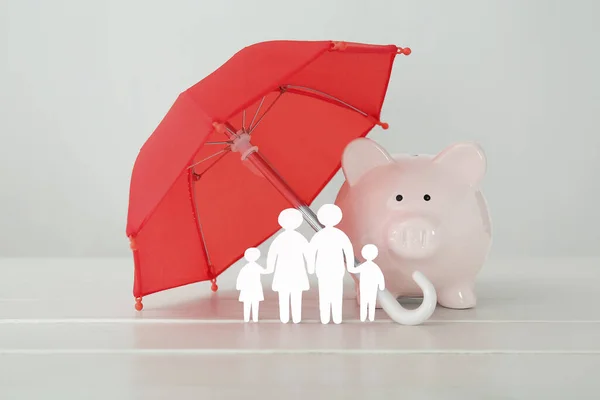 Figures of family with umbrella and piggy bank on white wooden table. Insurance concept
