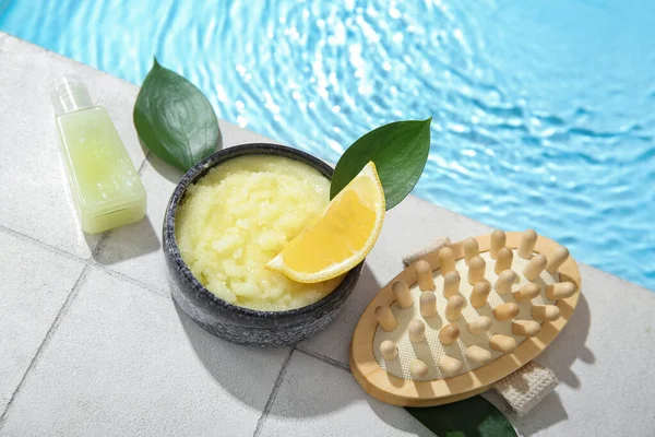Lemon body scrub with massage brush and cosmetic bottle on edge of swimming pool