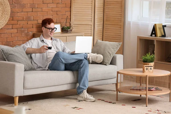Bonito Jovem Com Cartão Crédito Laptop Moderno Sentado Sofá Enquanto — Fotografia de Stock