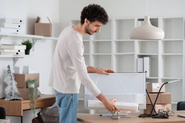 Genç adam ofiste streç filmle bilgisayar ekranını paketliyor.