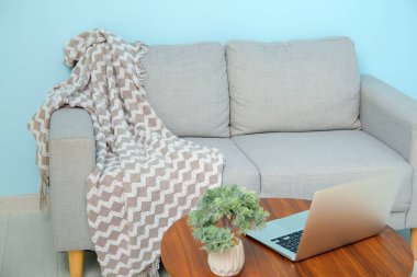 Mavi duvarın yanındaki sehpanın üzerinde rahat battaniyeli ve laptoplu gri kanepe.