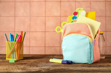 Renkli okul çantası, içinde not defterleri, suluboya resimler ve bej kiremit duvarın yanındaki ahşap masa üzerinde bir fincan işaretleyici.