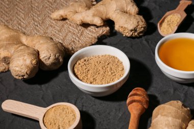 Taze zencefil kökleri, kase ve siyah zemin üzerinde kurumuş toz bulunan ahşap kaşık.