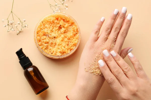 Female hands with body scrub and bottle of essential oil on color background, closeup