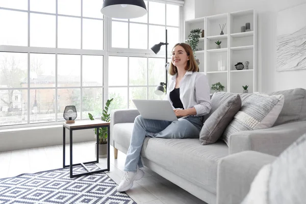 stock image Pretty young woman sitting on grey sofa and using modern laptop in light living room