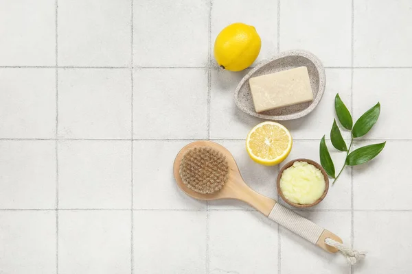 Bowl of lemon body scrub with massage brush and soap on white tile background