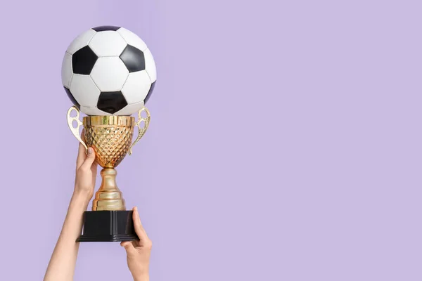 ライラックのバックグラウンドにゴールドカップとサッカーボールを持つ女性の手 — ストック写真