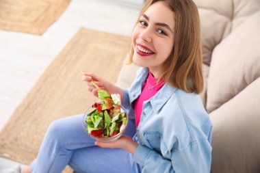 Evdeki kanepede oturan bir kase sebze salatalı genç kadın.