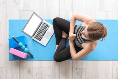 Dizüstü bilgisayarlı genç bir kadın spor salonunda yoga yapıyor.