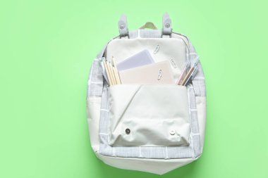 Yeşil arka planda farklı kırtasiye malzemesi olan gri okul çantası.