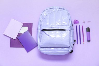 Mor arka planda farklı kırtasiye malzemesi olan leylak rengi okul çantası.