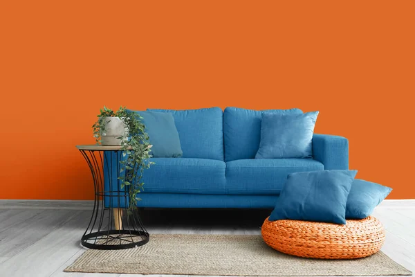 Interior of living room with blue sofa, pillows, table and houseplant near orange wall