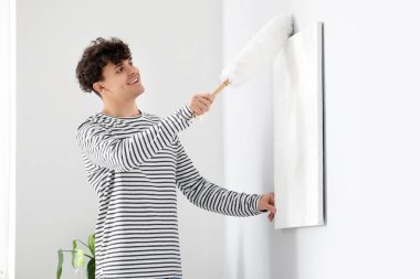 Genç adam yatak odasında toz bezi ile resim temizliyor.