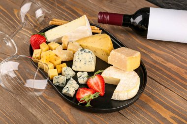 Lezzetli peynir parçaları, bardaklar ve şarap şişesi ahşap arka planda.