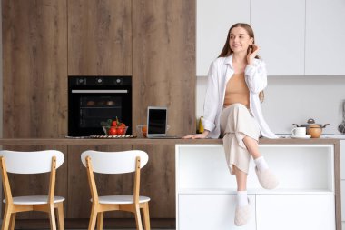 Mutfakta modern dizüstü bilgisayarla masada oturan mutlu genç bir kadın.