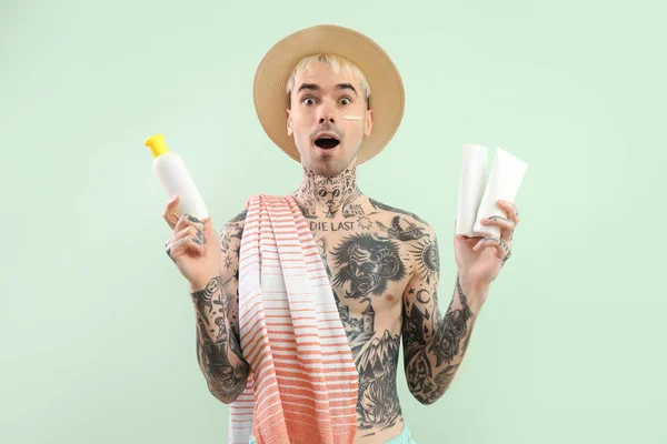 Surprised tattooed man with sunscreen creams on light green background