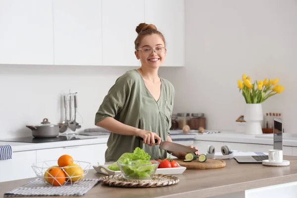 stock image Beautiful young woman cutting cucumber at table with modern laptop in light kitchen