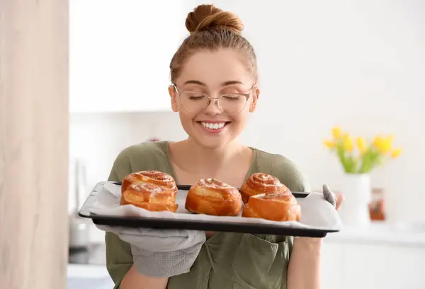 stock image Pretty young woman holding tray with fresh cinnamon rolls in kitchen