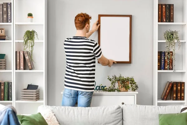 stock image Young redhead man hanging blank frame on light wall at home, back view