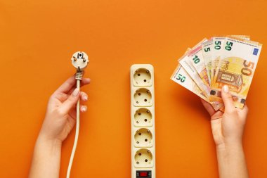 Turuncu arka planda parası, fişi ve prizi olan bir kadın. Elektrik faturası kavramı