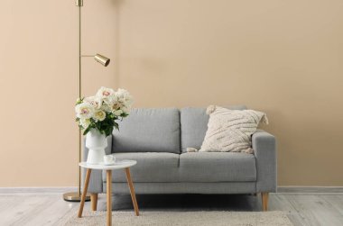 Kahve masası, kanepe ve bej duvarın yanında yer lambası olan beyaz şakayık vazoları.