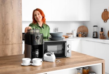 Modern mutfakta kahve makinesi, mikrodalga fırın ve mikseri olan genç kızıl saçlı kadın.